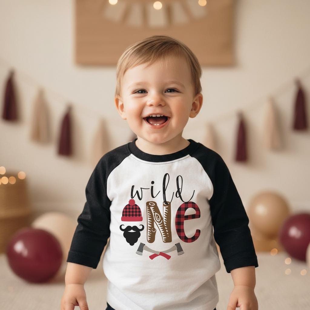 Wild One lumberjack birthday raglan tee with black sleeves, buffalo plaid beanie, beard graphic, and axes. Displayed in a nature-themed birthday setup with small pine trees. Ideal for lumberjack first birthday party, woodland 1st birthday outfit, and rustic Wild One celebrations.