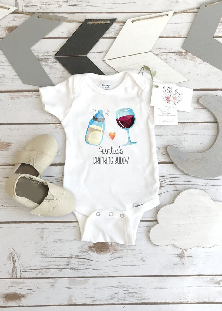 May include: A white baby onesie with a graphic of a baby bottle and a glass of wine. The text on the onesie reads 'Auntie's Drinking Buddy'. The onesie is on a wooden surface with two pairs of baby shoes and a wooden sign that says 'bella lei'.