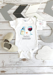 May include: A white baby onesie with a graphic of a baby bottle and a glass of wine. The text on the onesie reads 'Auntie's Drinking Buddy'. The onesie is on a wooden surface with two pairs of baby shoes and a wooden sign that says 'bella lei'.