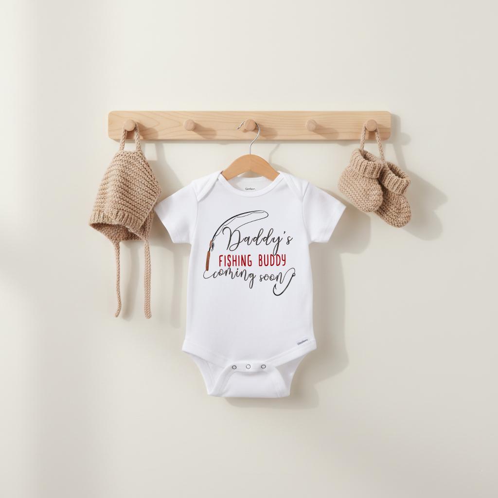 White onesie hanging on wood rack with knit bonnet and booties, Daddy’s Fishing Buddy Coming Soon baby reveal outfit, fishing themed pregnancy reveal gift for expecting dad, baby shower gift for outdoorsy families, cute new baby announcement outfit for photos or surprise reveal videos.